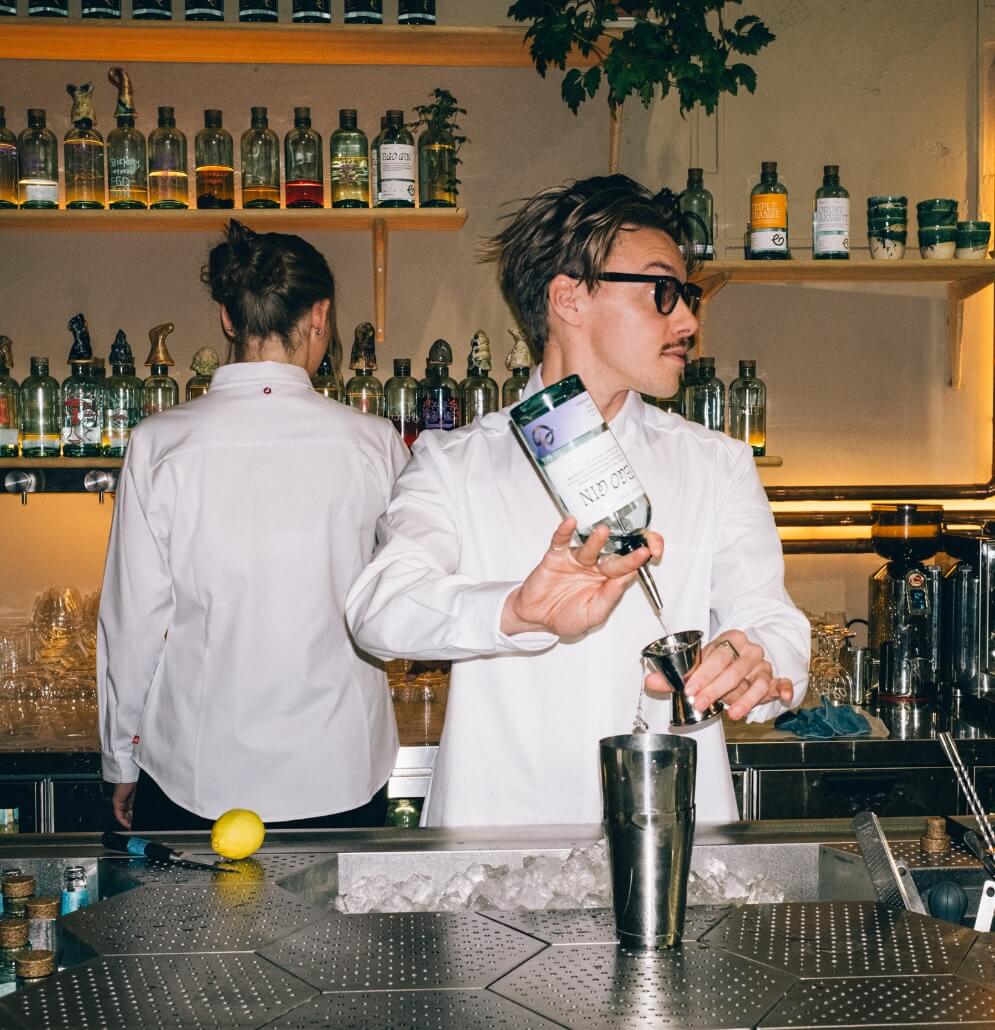 Bartender häller gin i shaker bakom bardisk med flaskor i bakgrunden.