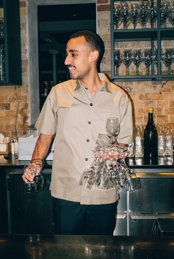 Man som arbetar bakom en bar och bär beige arbetskläder. Håller flera vinglas i handen, med barutrustning och flaskor i bakgrunden.