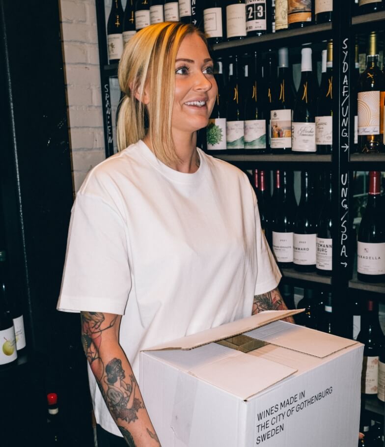 Kvinna i oversize t-shirt bär en kartong med vinflaskor i ett vinrum på en restaurang.