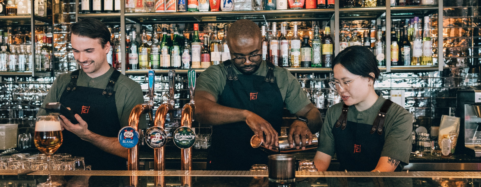Tre bartendrar bakom en bardisk, klädda i gröna kockskjortor och svarta förkläden och i bakgrunden syns flera flaskor. 